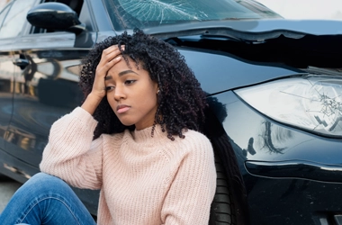 A woman holding her head while sitting next to a car after a car accident.